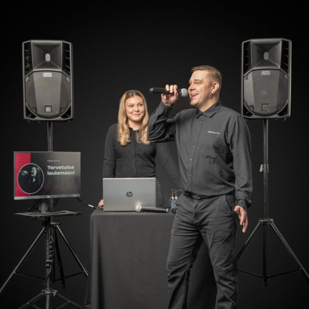 Kaksi henkilöä Popmasterin DJ- ja karaokepalvelun laitteiston äärellä: toinen laulaa mikrofoniin, toinen käyttää tietokonetta. Kuvassa näkyvät kaksi kaiutinta, laulunäyttö, mikrofoni ja pöydälle asetettu kannettava tietokone.